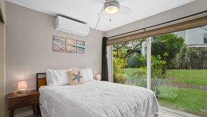 A bedroom with a large bed, a nightstand with a lamp, a ceiling fan, and a view of a garden through a sliding glass door. - Cozy Modern Kamahana Studio, Pool, WiFi (Princeville)
