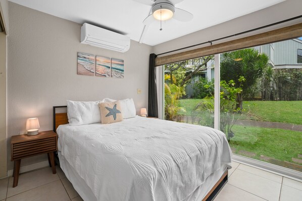A bedroom with a large bed, a nightstand with a lamp, a ceiling fan, and a view of a garden through a sliding glass door.