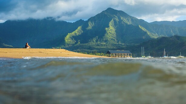 A beach with a person sitting on the sand, a small building, and mountains in the background. - Cozy Modern Kamahana Studio, Pool, WiFi (Princeville)