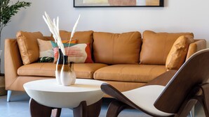 A modern living room with a brown leather sofa, a white coffee table, and a brown chair. There is a painting on the wall and a plant on the left.