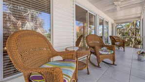 A covered patio with wicker furniture and a glass table, overlooking a garden with palm trees.