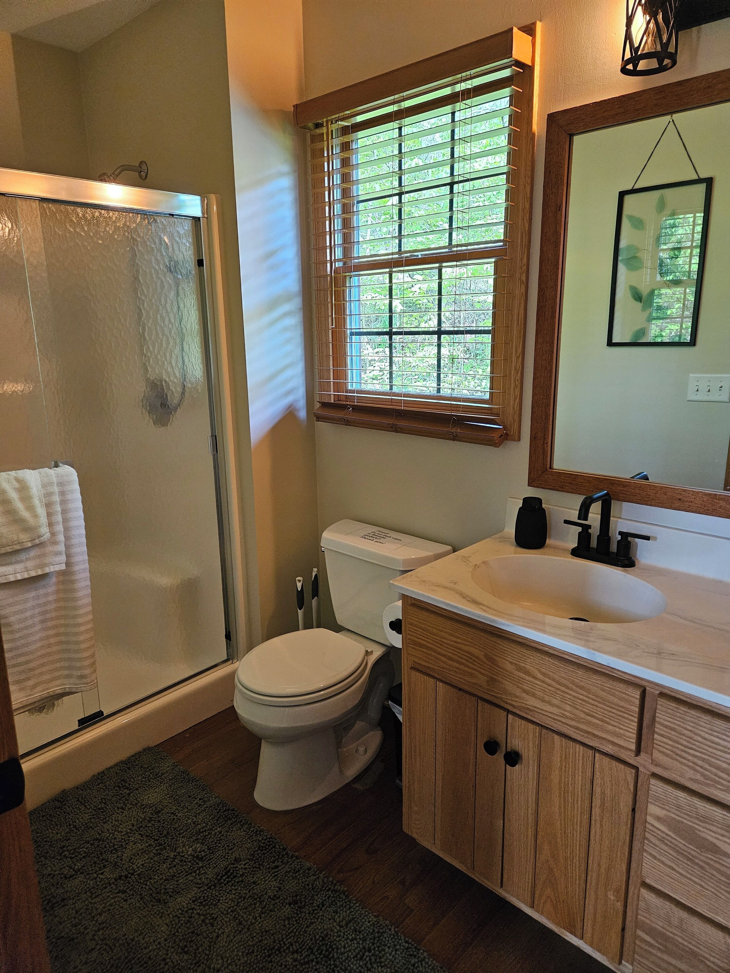 A bathroom with a shower, toilet, sink, and window with blinds.