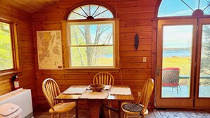 A wooden cabin interior with a dining table, four chairs, a map on the wall, and a view of the outdoors through a large window.