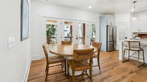 A modern kitchen with a central island, wooden flooring, and a dining area with wicker chairs. - 4 Elderberry Ln, New Build, Elevator, Steps to Beach, Putting Green, Private Pool (Hilton Head Island)