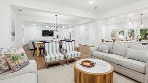 A spacious living room with a large sofa, striped chairs, a round coffee table, and a dining area with a TV and modern decor. - 4 Elderberry Ln, New Build, Elevator, Steps to Beach, Putting Green, Private Pool (Hilton Head Island)