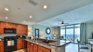 Une cuisine spacieuse dotée de plans de travail en granit, d’armoires en bois et d’un grand îlot central. Elle comprend un micro-ondes, une cuisinière et un évier encastrés. La cuisine donne sur un balcon offrant une vue sur l’océan.