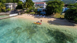 Ein Strand mit klarem türkisblauem Wasser, ein weißes Gebäude mit rotem Dach und ein großer Baum.