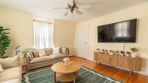 Una sala de estar con un sofá, una mesa de centro con un florero, un televisor en la pared y un aparador de madera con plantas.