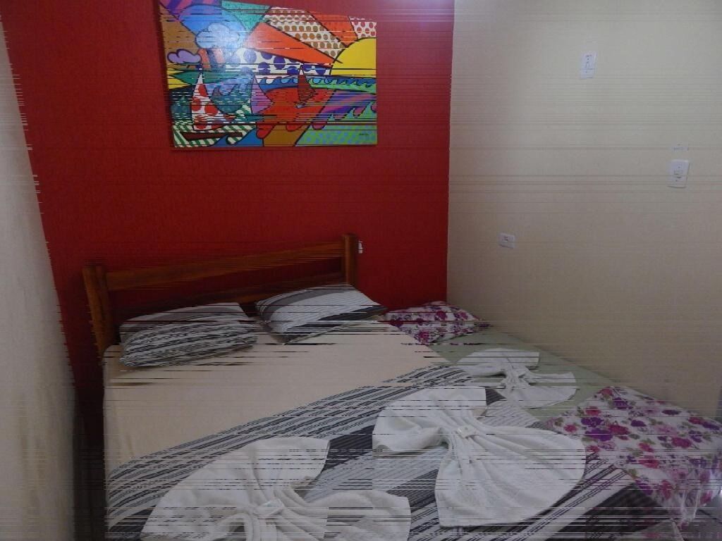 A small, narrow hotel room with a single bed, a colorful painting on the wall, and a red accent wall.
