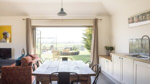 A modern living room with a sofa, TV, and dining table. There is a kitchen area with a sink and cabinets. - Stylish cottages, countryside views, wood burning hot tubs, 20 mins to Cambridge (Royston)