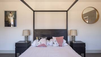 A modern bedroom with a large bed, two bedside tables with lamps, a dark headboard, a round mirror, and a framed picture on the wall.