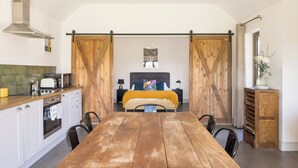 A kitchen with a wooden table and chairs, a bed with a yellow blanket, and a sliding barn door. - Stylish cottage, countryside views, wood burning hot tub, 20 mins to Cambridge (Royston)