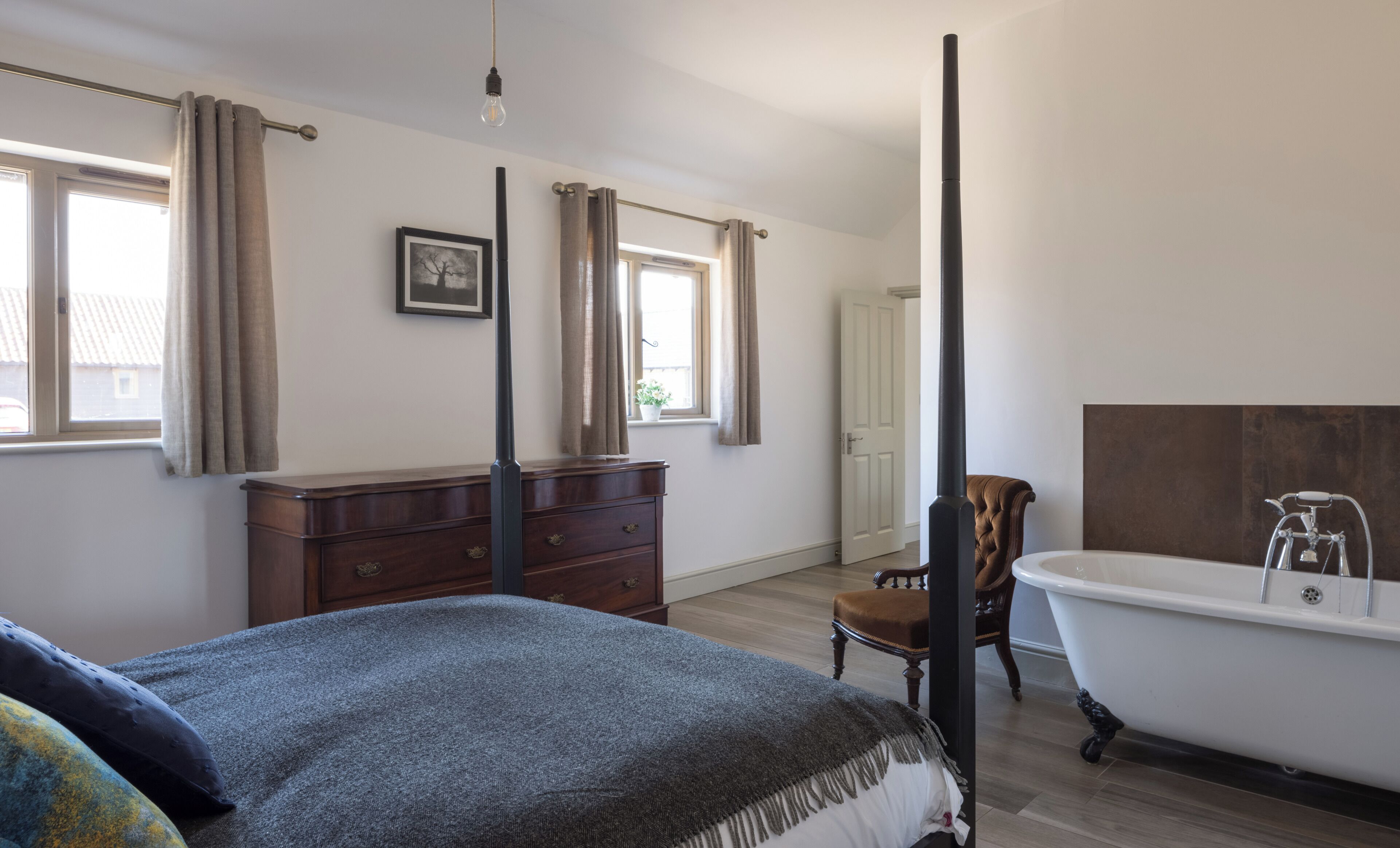 A bedroom with a bed, a dresser, a chair, a window with curtains, a picture on the wall, and a bathtub.