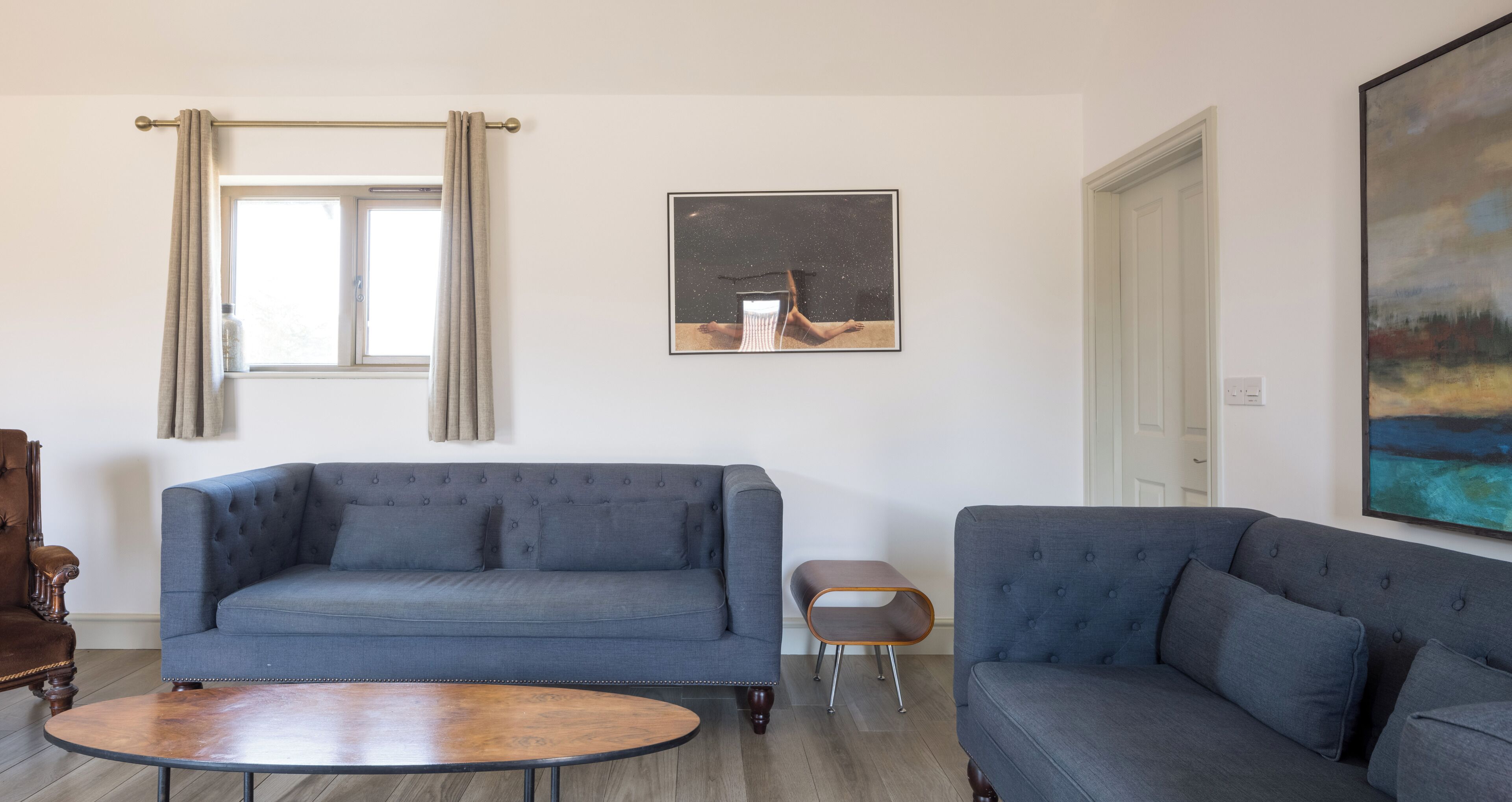 A living room with two sofas, a coffee table, a side table, a window with curtains, a framed picture, and a door.
