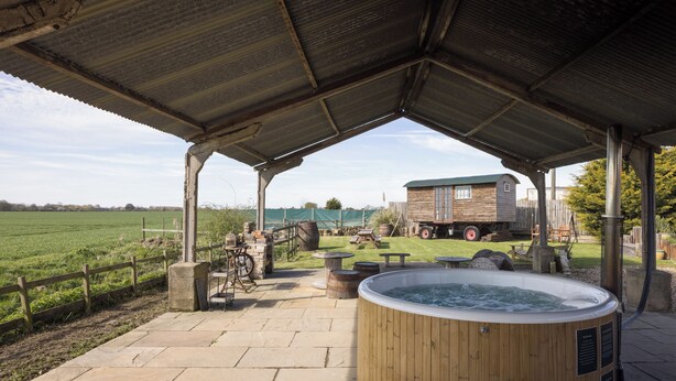 A rustic outdoor patio with a hot tub, wooden barrels, and a wooden structure in the background. - Stylish cottages, countryside views, wood burning hot tubs, 20 mins to Cambridge (Royston)