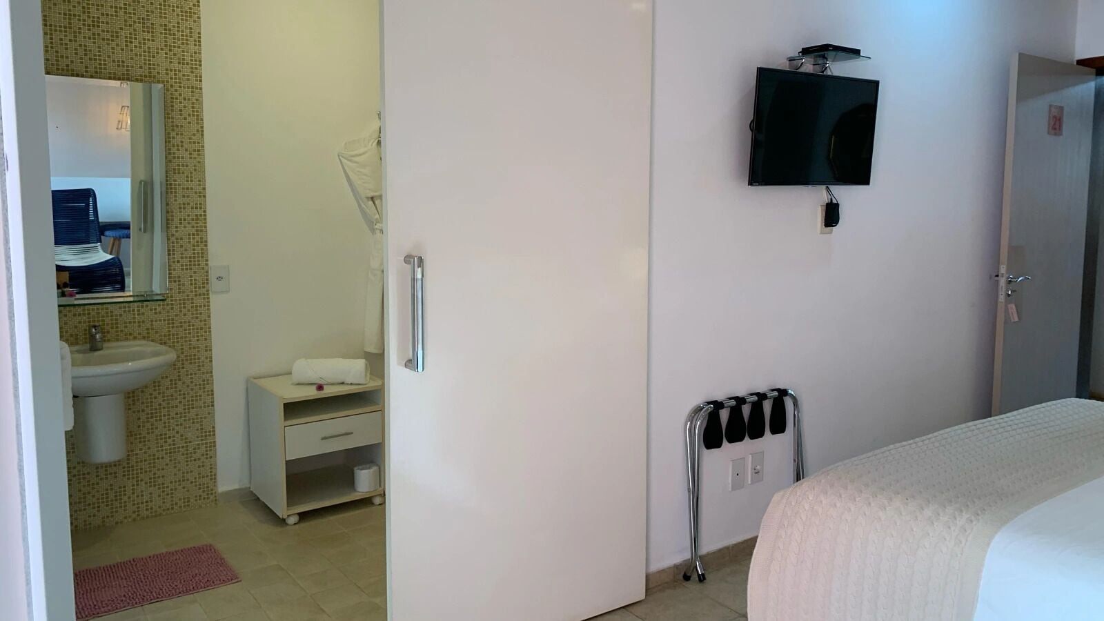 Quarto com cama, televisão presa na parede, radiador e banheiro visível por uma porta aberta.
