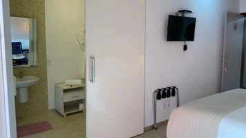 Quarto com cama, televisão presa na parede, radiador e banheiro visível por uma porta aberta.