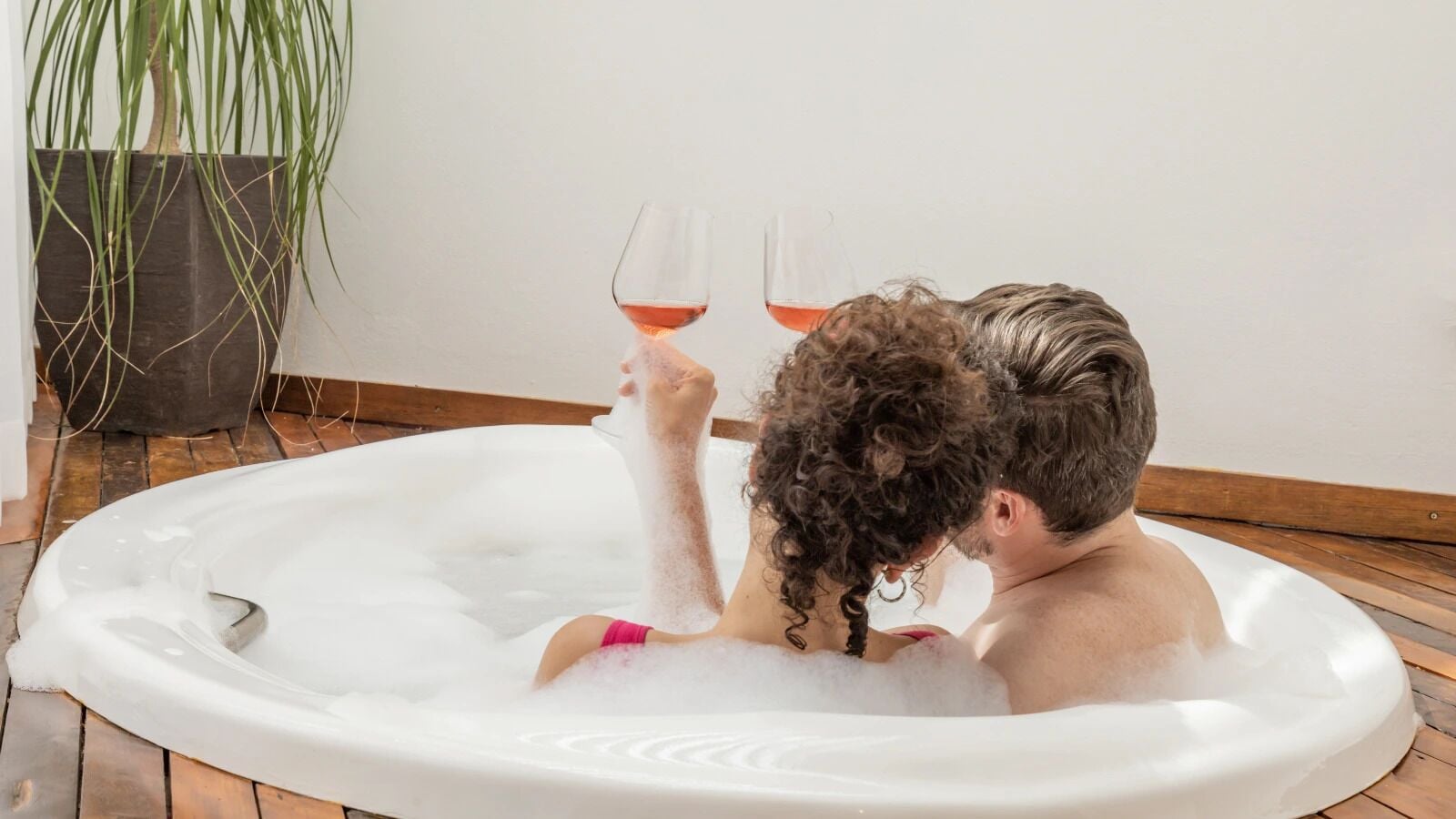 A couple in a hot tub with a glass of wine, surrounded by a wooden floor and a potted plant.