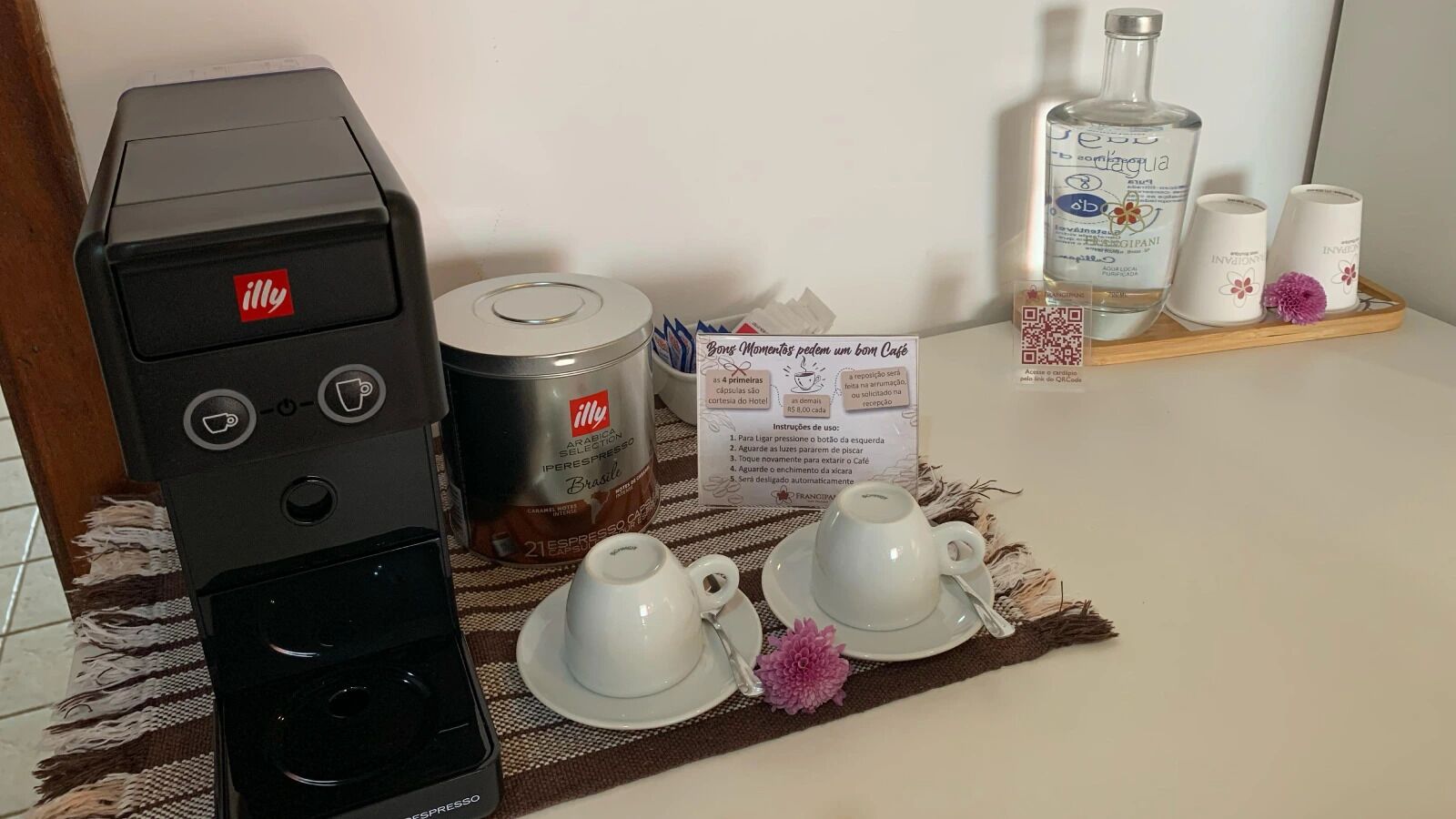 A coffee machine, a coffee pod, a cup, a saucer, a tray with a bottle and two candles, and a sign with instructions.