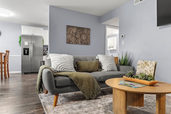 A modern living room with a grey sofa, a wooden coffee table, and a kitchen area in the background.
