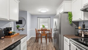 A modern kitchen with white cabinets, a granite countertop, a stainless steel refrigerator, and a dining area with wooden chairs and a table.