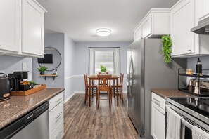 Una cocina moderna con armarios blancos, encimera de granito, refrigerador de acero inoxidable y un comedor con sillas de madera y una mesa.