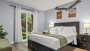 A bedroom with a bed, bedside tables, a nightstand, a window with curtains, and a view of greenery outside.