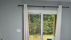 A bedroom with a bed, a small table, a chair, a plant, and a sliding glass door leading to an outdoor area.