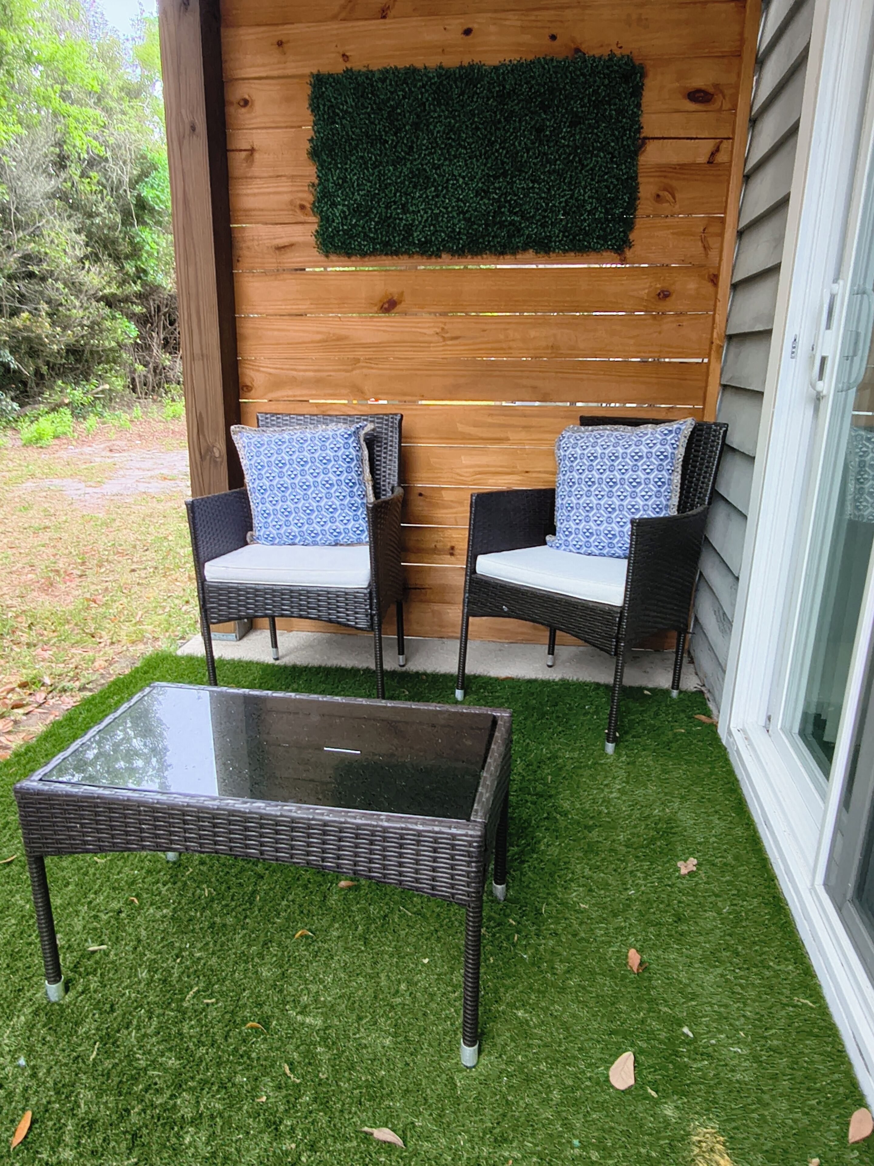 En terrasse med to fletstole, et glasbord og en trævæg med grønt kunstgræs.