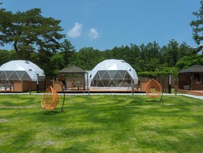Trois structures en forme de dôme géodésique, dotées de terrasses en bois attenantes, et entourées d’une zone herbeuse et d’arbres.