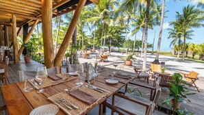 An outdoor dining area with wooden tables and chairs, surrounded by palm trees and a clear view of the ocean. - NANNAI Milagres (Passo de Camaragibe)