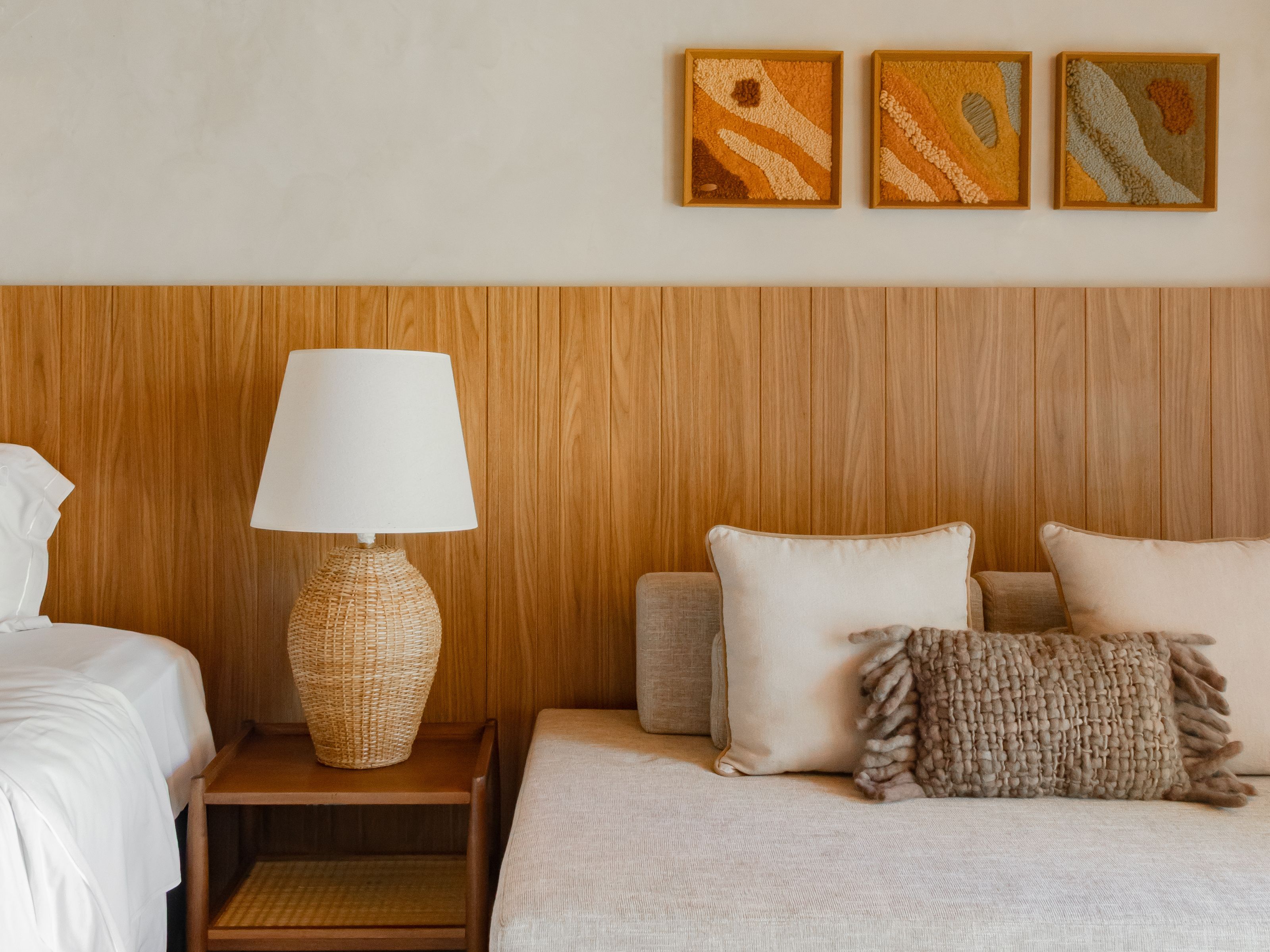 Un dormitorio con cabecera de madera, mesita de noche con lámpara, cama con almohadas y tres cuadros enmarcados en la pared.