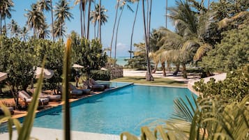 A pool area with lounge chairs and palm trees, surrounded by greenery and a view of the ocean.