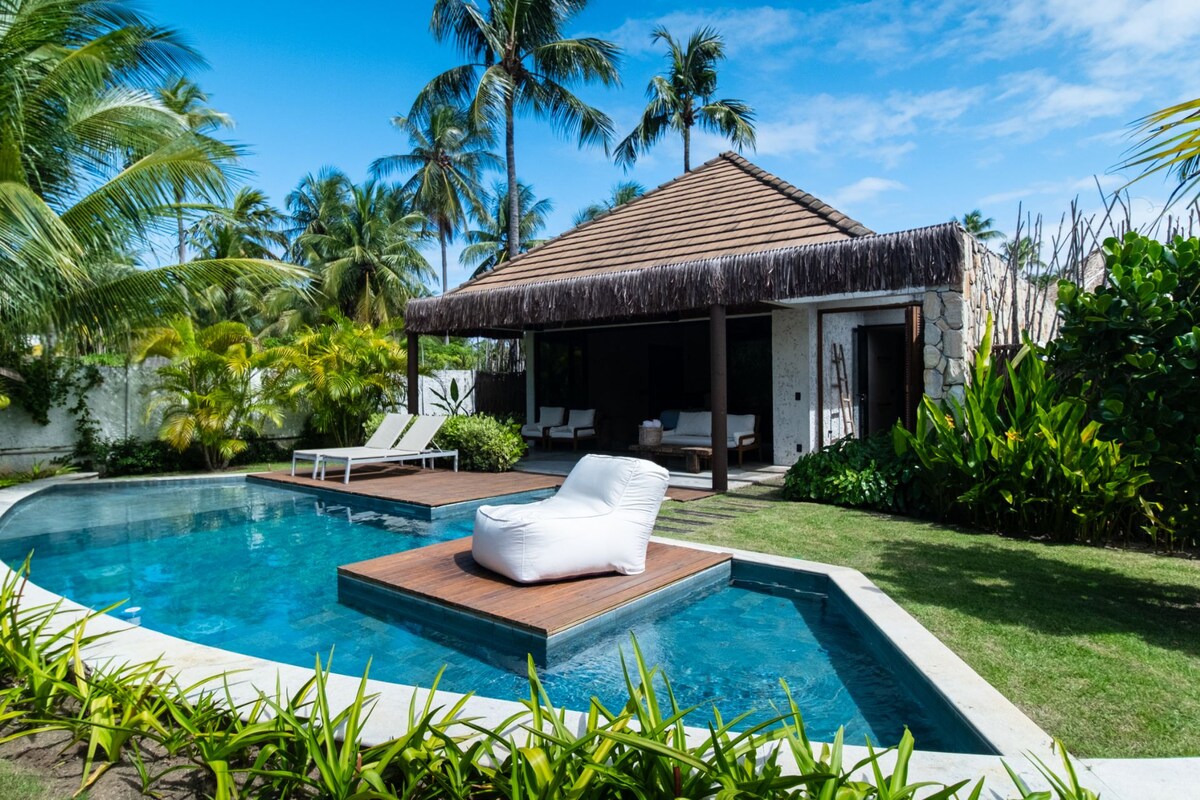 A swimming pool with a lounge chair, a thatched-roof structure, and lush greenery.