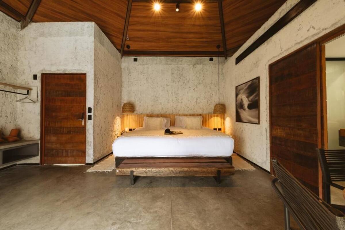 Un dormitorio con una cama de madera, un banco, una puerta y una lámpara fijada a la pared.