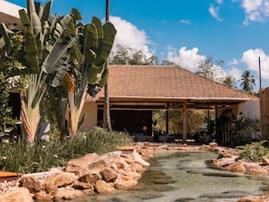 A tropical resort with a thatched-roof pavilion, a clear water feature, and lush vegetation.