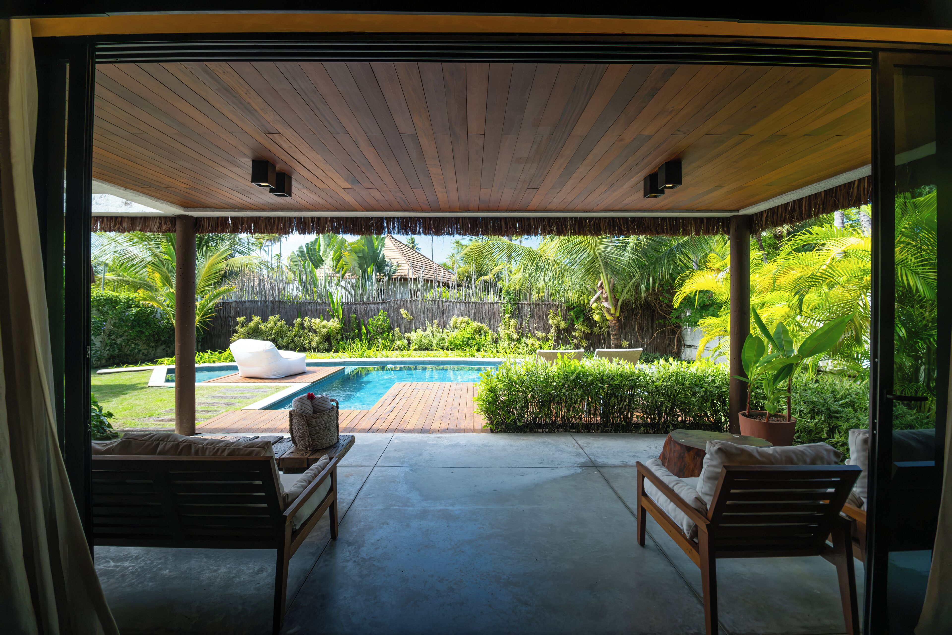 A covered outdoor area with a pool, lounge chairs, and a view of a tropical garden.