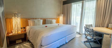 A modern hotel room with a large bed, a wooden headboard, a bedside table with a lamp, and a dining area with a table and chairs.