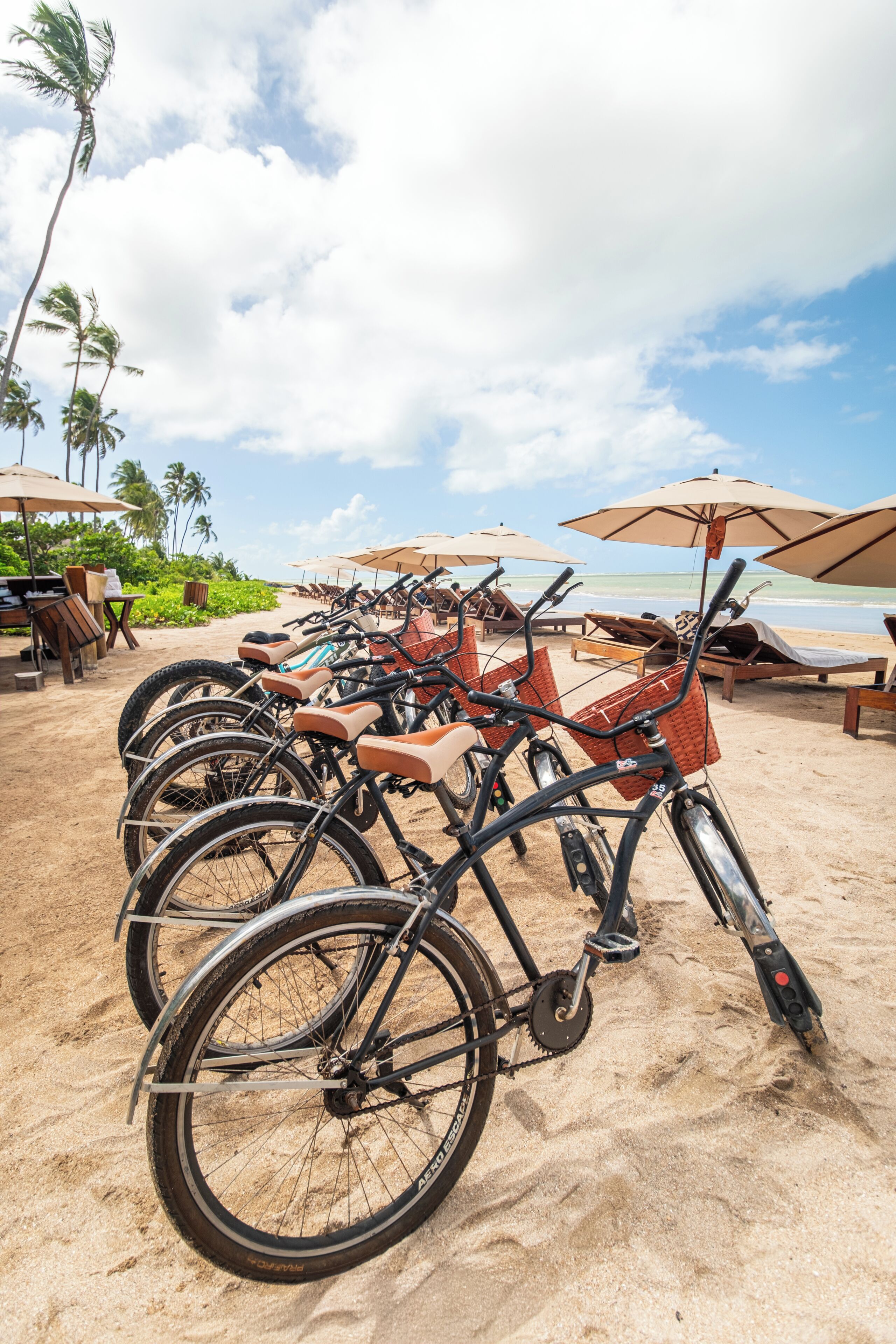 Des vélos sont garés sur une plage de sable, avec des parasols et des chaises longues en arrière-plan.