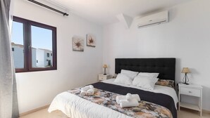 A bedroom with a large bed, two bedside tables, an air conditioning unit, and a window with a view of buildings.