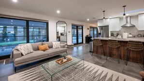 A modern kitchen with a bar area, large windows, and a living room with a sofa and coffee table. - Modern Desert Oasis - Shuffleboard, Hiking, Pool & Putting green! (Phoenix)