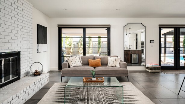 A modern living room with a white brick fireplace, a glass coffee table, a grey sofa with cushions, and a view of the outdoor pool area through large sliding glass doors. - Modern Desert Oasis - Shuffleboard, Hiking, Pool & Putting green! (Phoenix)