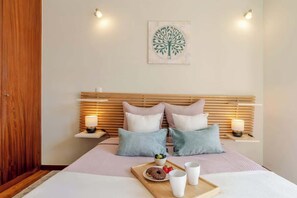 A neatly arranged bedroom with a wooden headboard, bedside tables with lamps, a bed with a pink and white bedspread, and a tray with a donut and fruits on the bed. - Bright & Airy Flat w/ Balcony by HostWise (Porto)