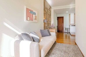 Uma sala de estar moderna com sofá, parede de destaque em pedra, cozinha ao fundo e corredor que dá para outro espaço.
