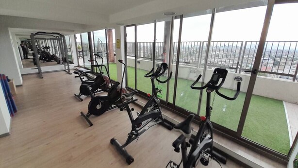 A gym with exercise bikes and cardio machines, large windows with a view of the city, and a balcony with a green lawn.