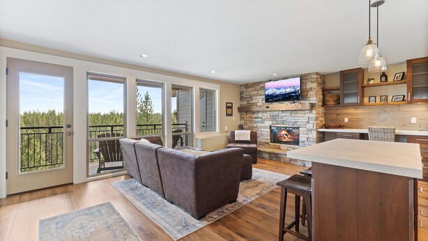 A spacious living area with a fireplace, a television mounted on a stone wall, and a balcony with a view of trees.
