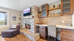 A cozy living room with a stone fireplace, a comfortable sofa, a dining area with wooden cabinets, and a television mounted above the fireplace.