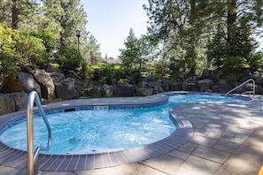 Un jacuzzi rodeado de rocas y árboles, con un borde de azulejos y una escalera metálica.
