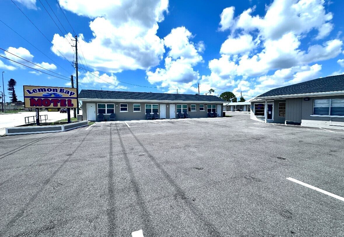 Motel Lemon Bay con un amplio estacionamiento, un edificio de una sola planta y un cielo azul despejado con nubes.
