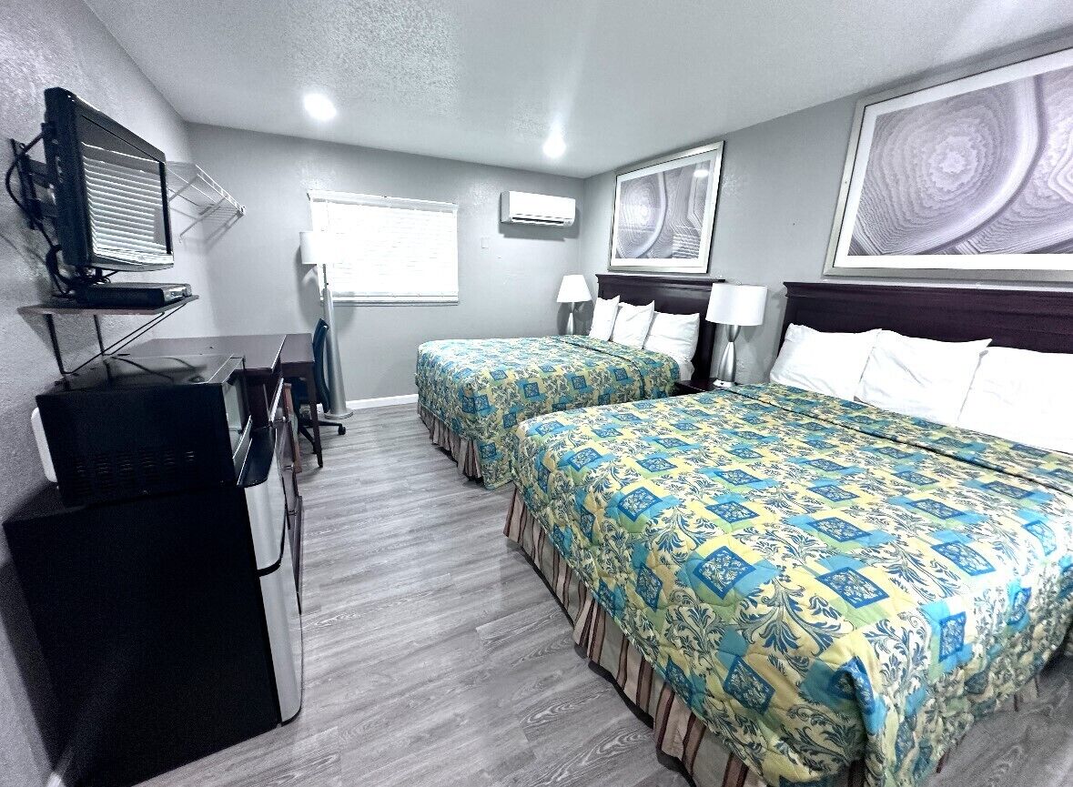 Habitación de hotel con dos camas, un escritorio con computadora, un televisor y obras de arte en las paredes.